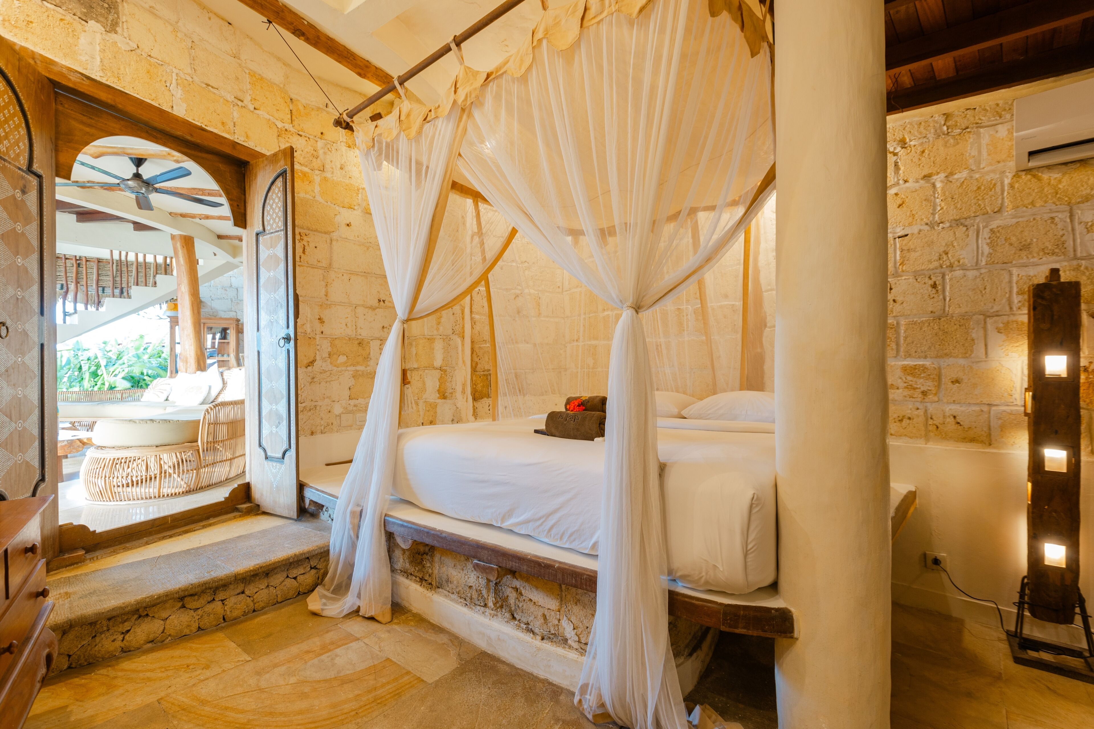 A room with a stone wall, a bed with white bedding, a canopy, and a view of a balcony with outdoor furniture.