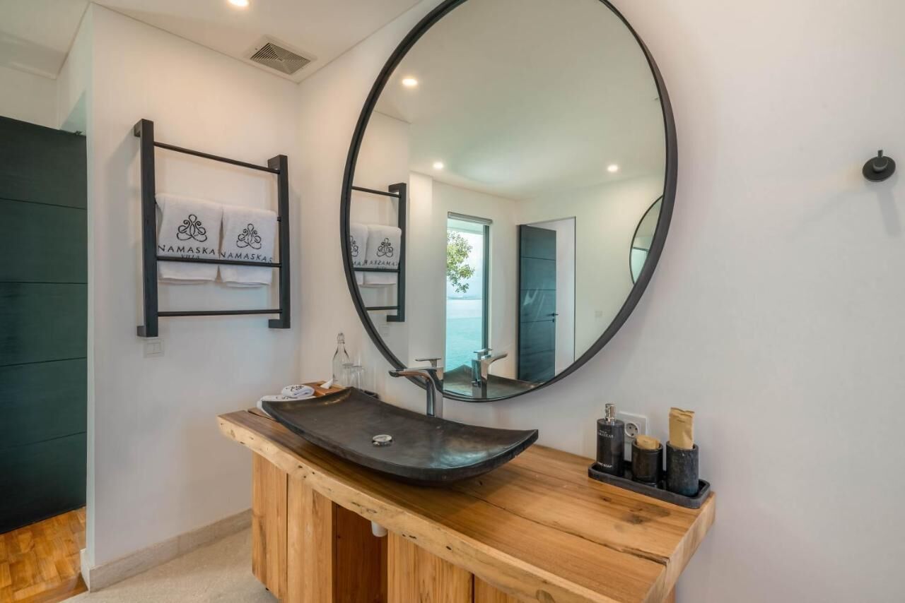 A modern bathroom with a wooden vanity, a large round mirror, and a towel rack with branded towels.