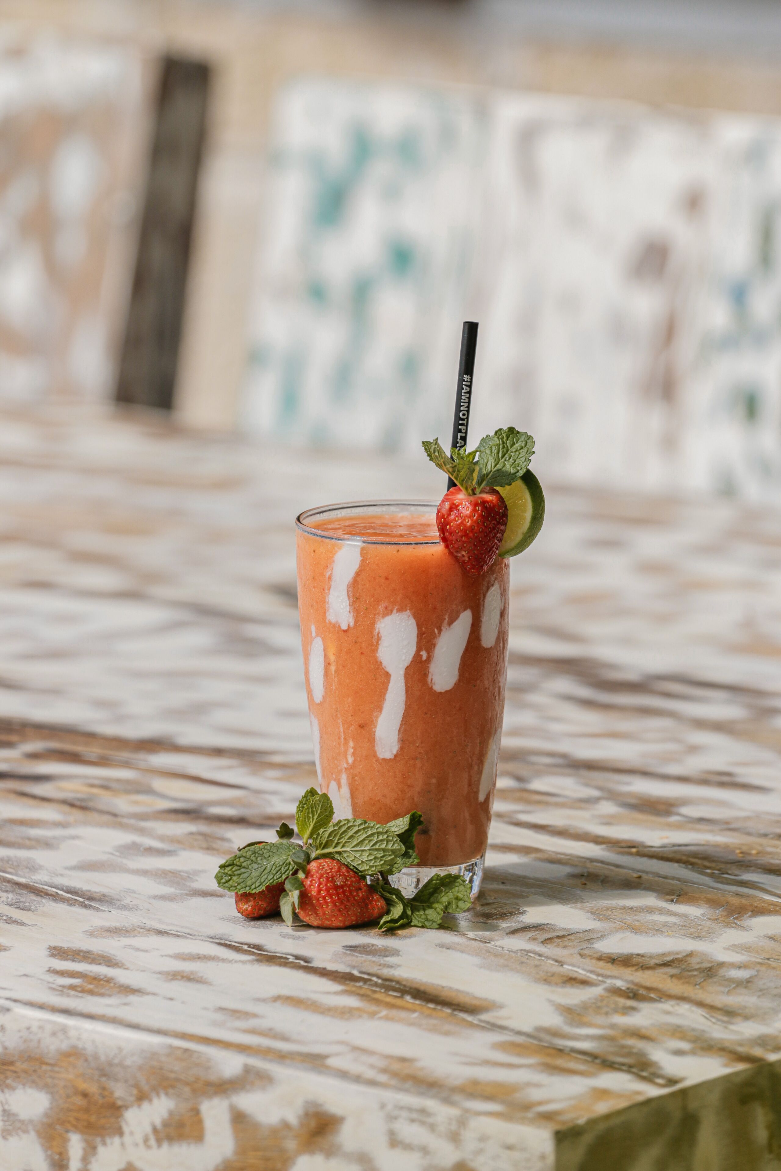Een hoog glas aardbeien smoothie met rietje, versierd met een aardbei, muntblaadjes en een partje limoen, op een tafel met patroon.