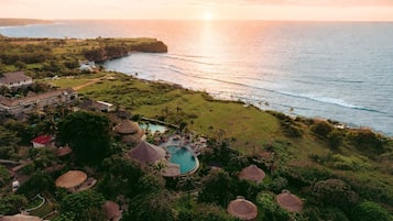 An aerial view of a resort with a swimming pool, thatched-roof structures, and a view of the ocean.
