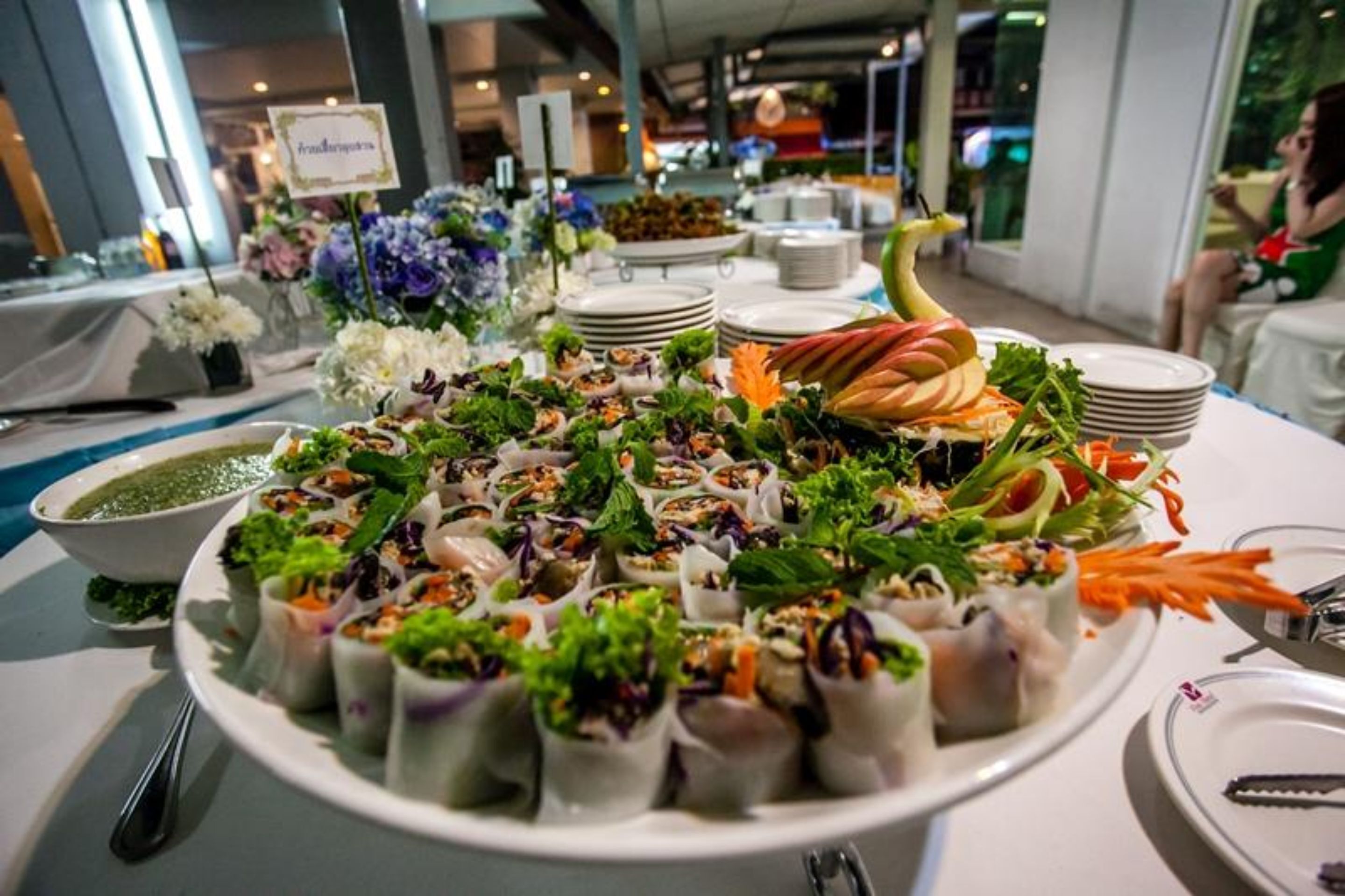 Uma mesa de buffet com diversos pratos, incluindo rolinhos primavera e saladas, em uma área de refeições com mesas e cadeiras.