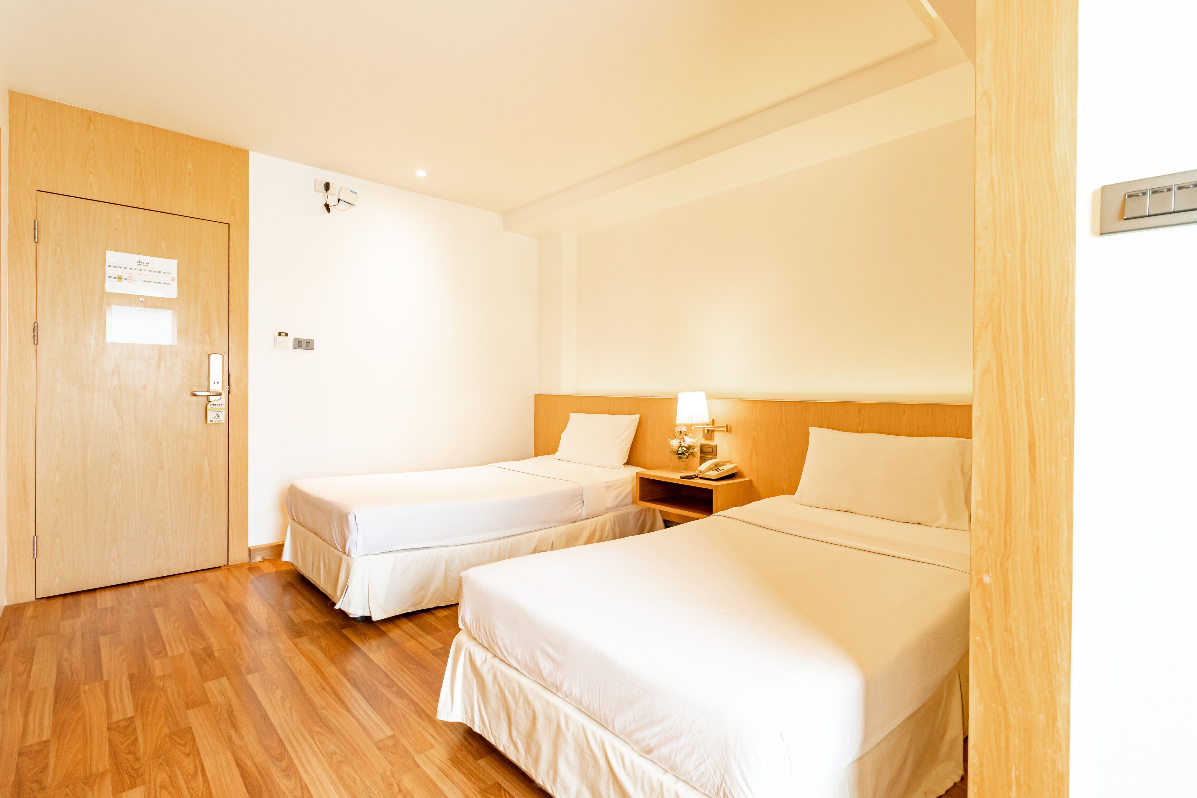A hotel room with two single beds, a wooden wardrobe, a bedside table with a lamp, and a wall-mounted thermostat.