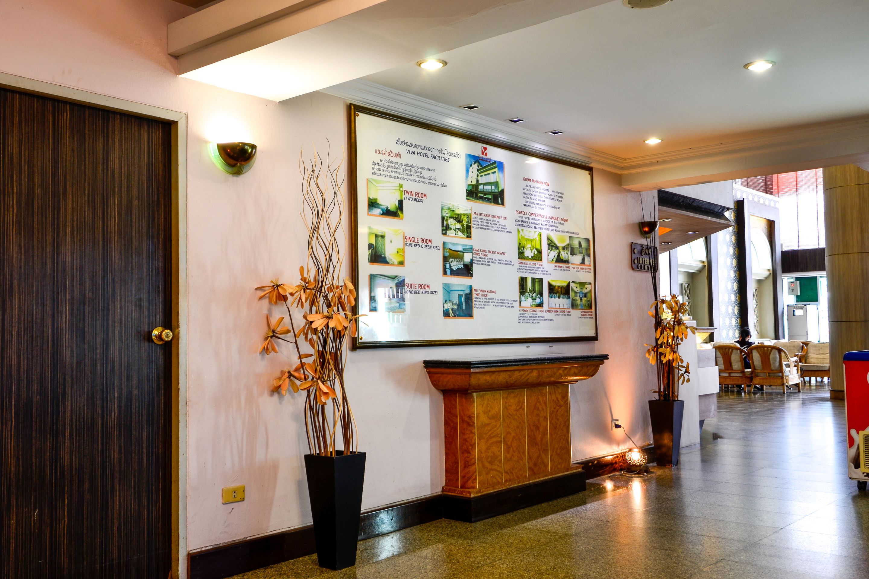 Een hotel receptie met een groot informatief poster, een houten receptiebalie en potplanten.