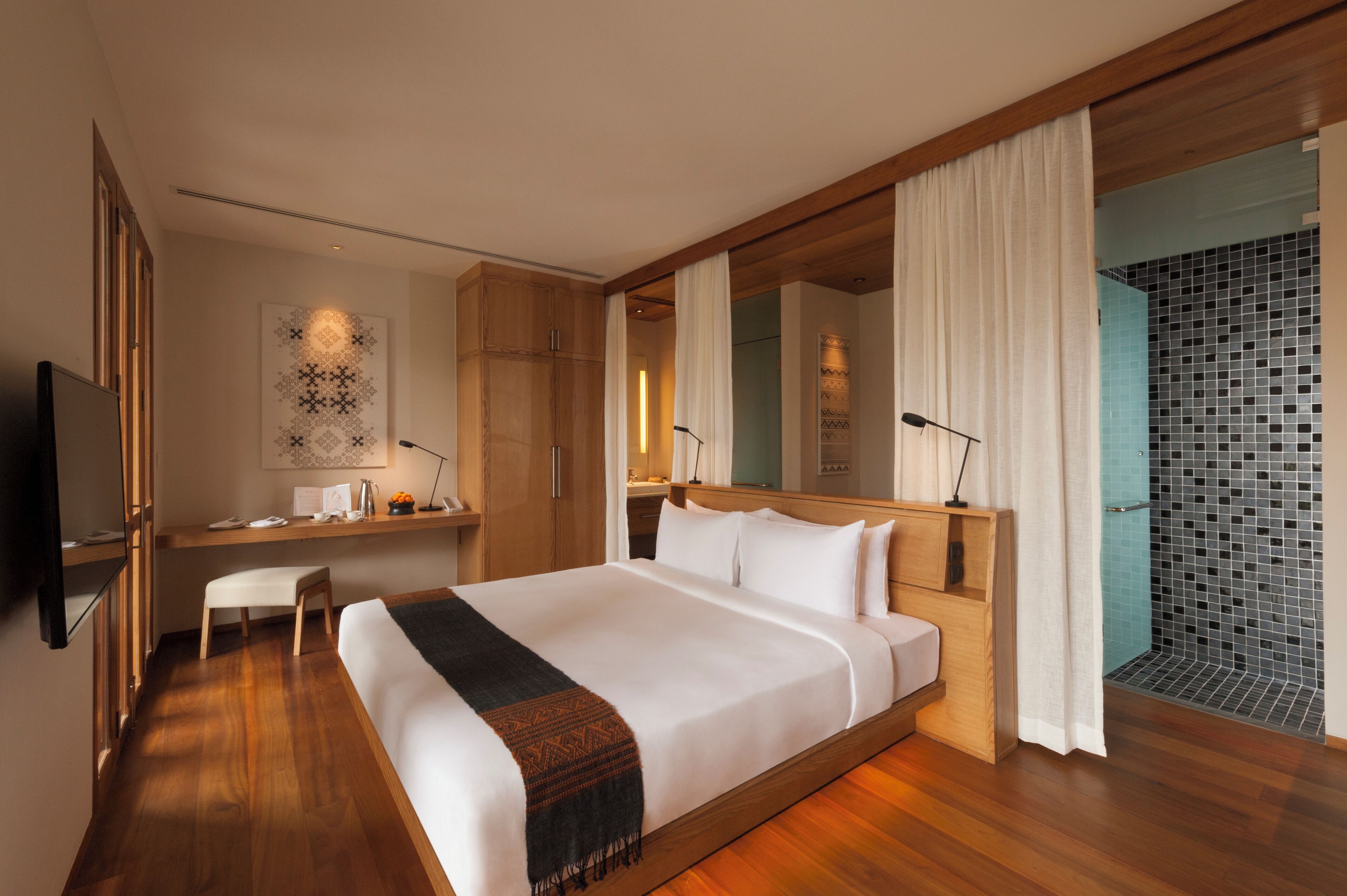 A modern hotel room with a large bed, wooden flooring, a desk with a chair, a TV, and a bathroom with a tiled shower.