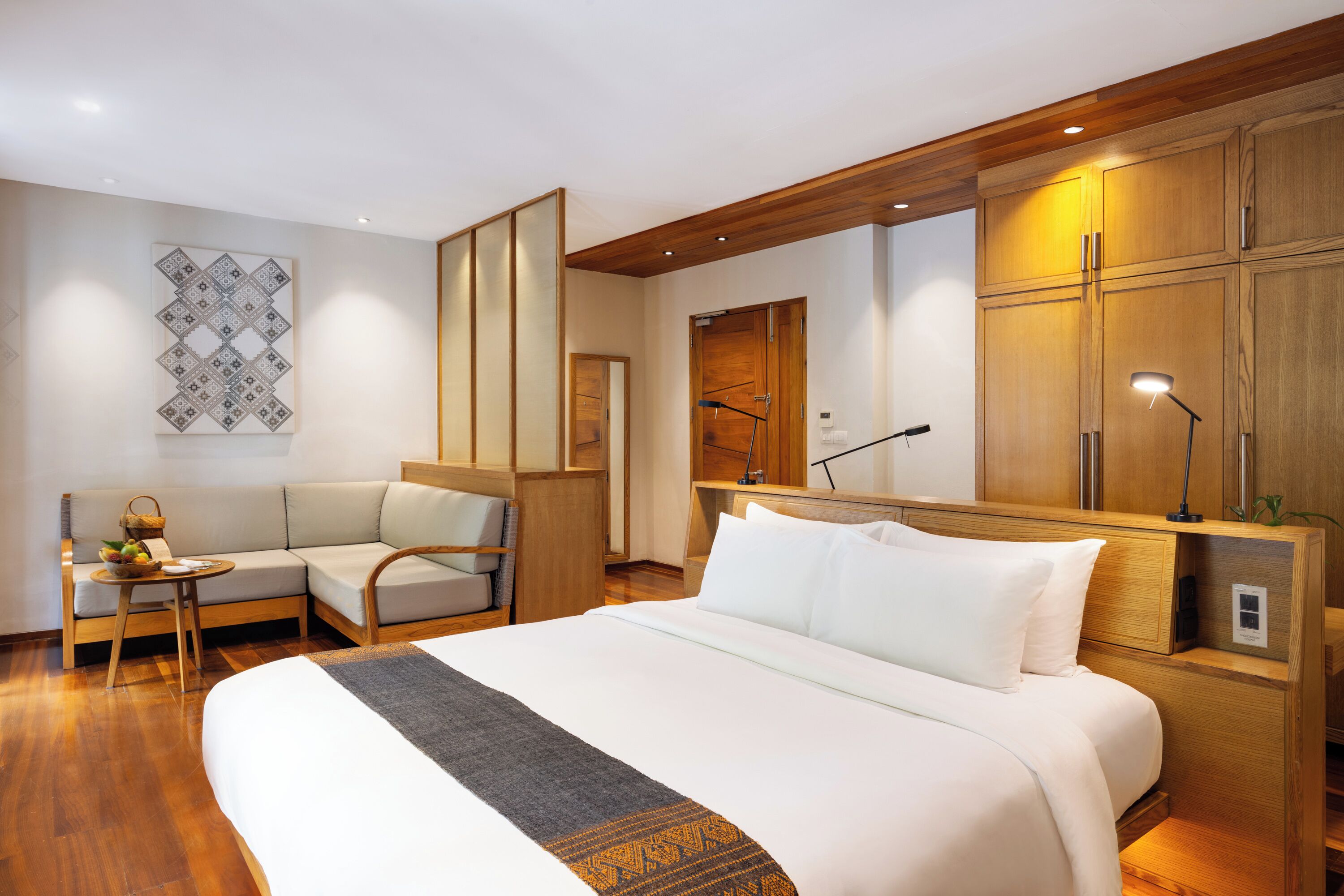 A hotel room with a large bed, a sofa, a small table with a fruit bowl, and wooden cabinets.
