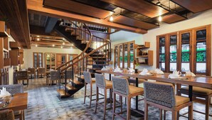 Un vaste espace dédié à la salle à manger, avec de grandes tables et des chaises en bois, un escalier central et de grandes fenêtres offrant une vue sur la verdure environnante.