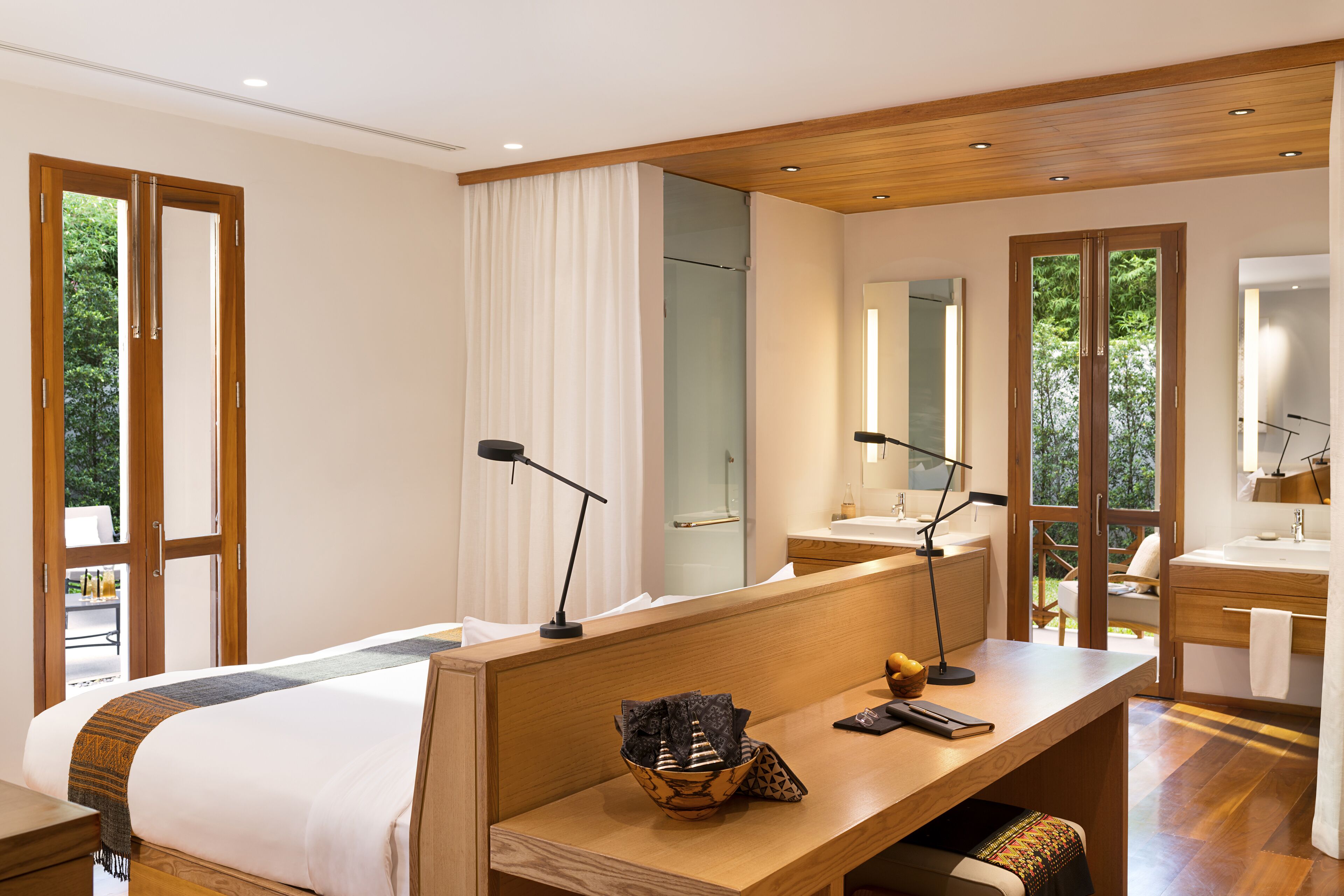 A modern hotel room with a large bed, a desk with a computer, a bathroom with a sink and mirror, and large windows with curtains.