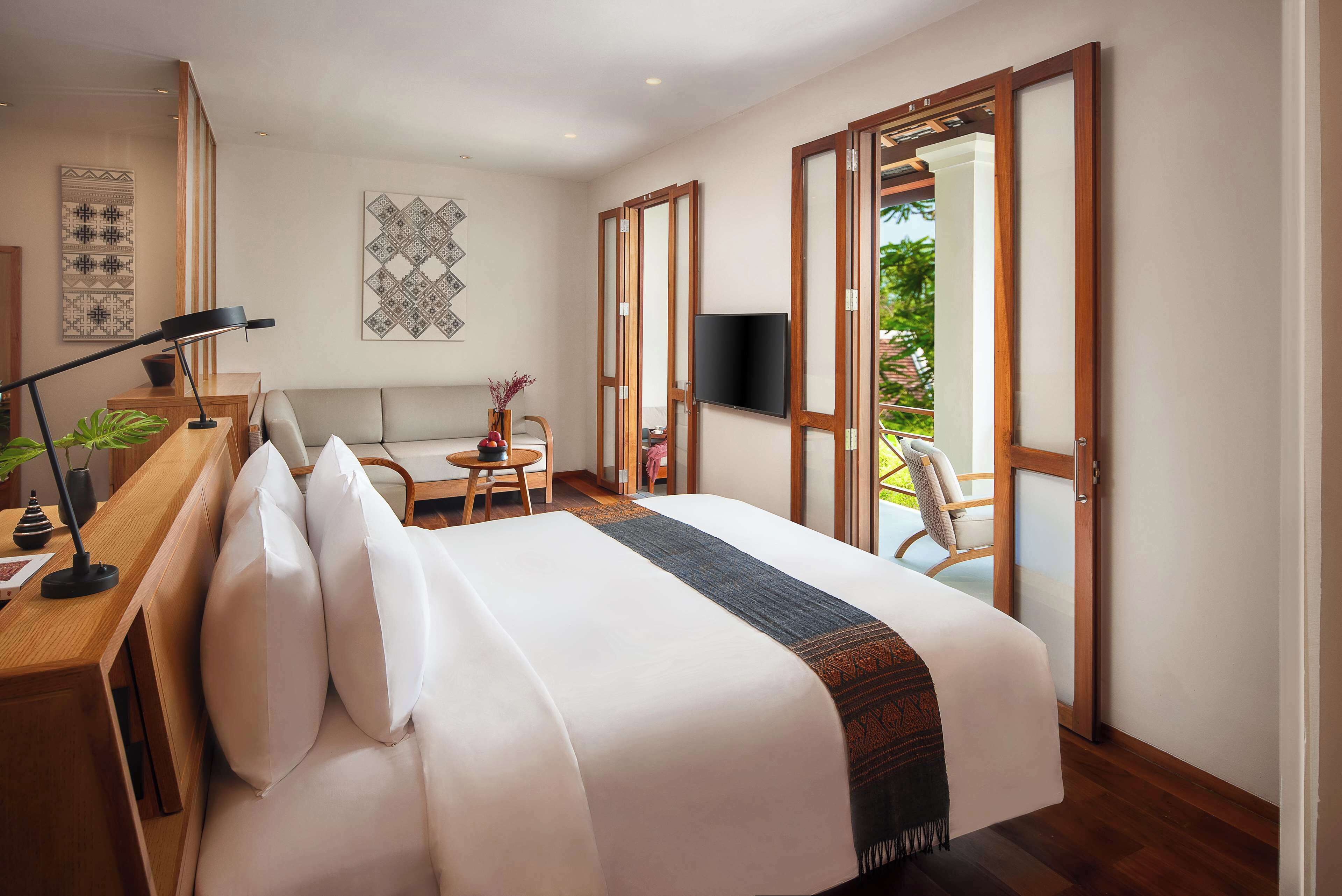 A hotel room with a bed, a desk, a chair, a TV, and a balcony with a view of greenery.