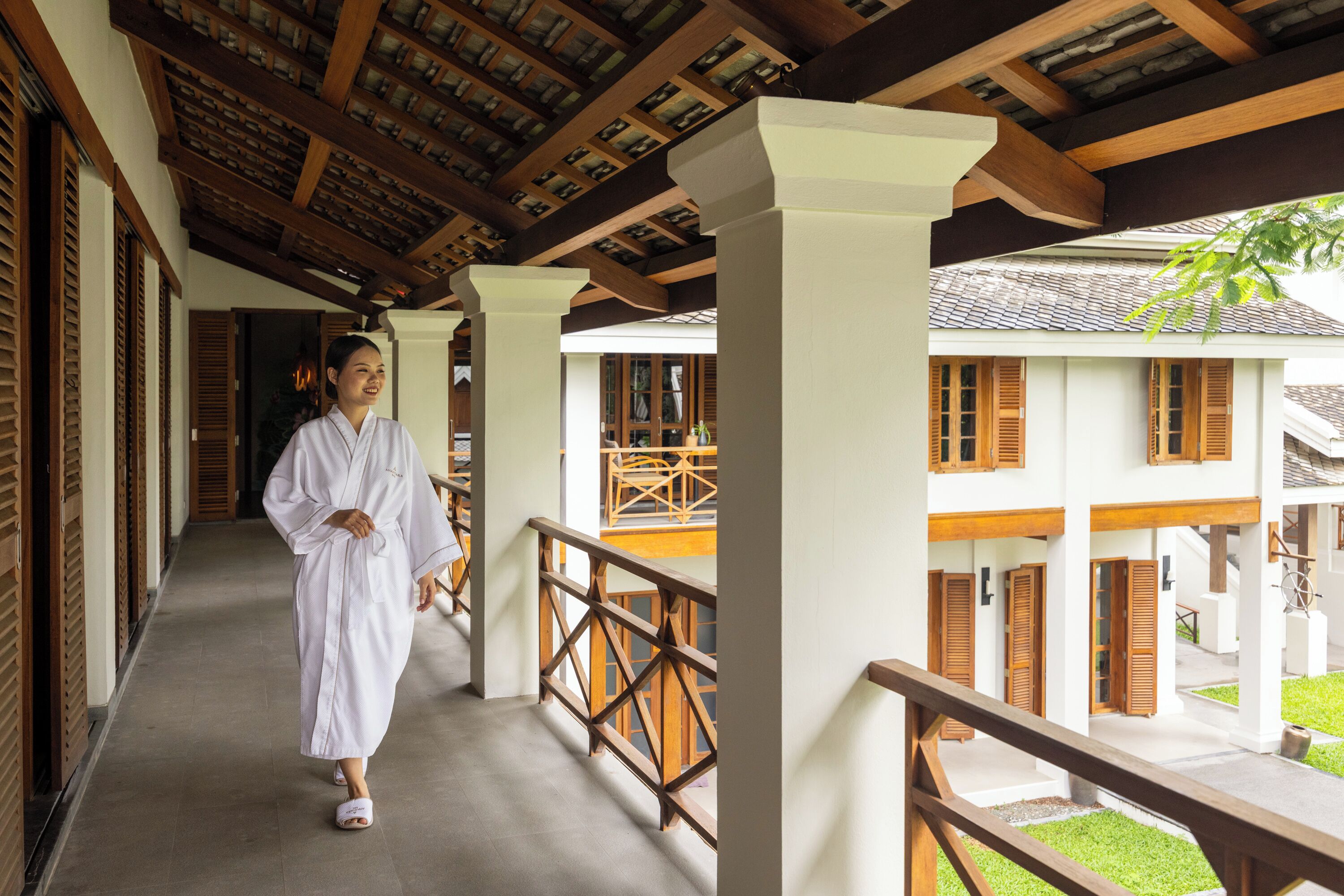 A corridor with wooden railings and a tiled floor, leading to a building with multiple windows and wooden shutters.