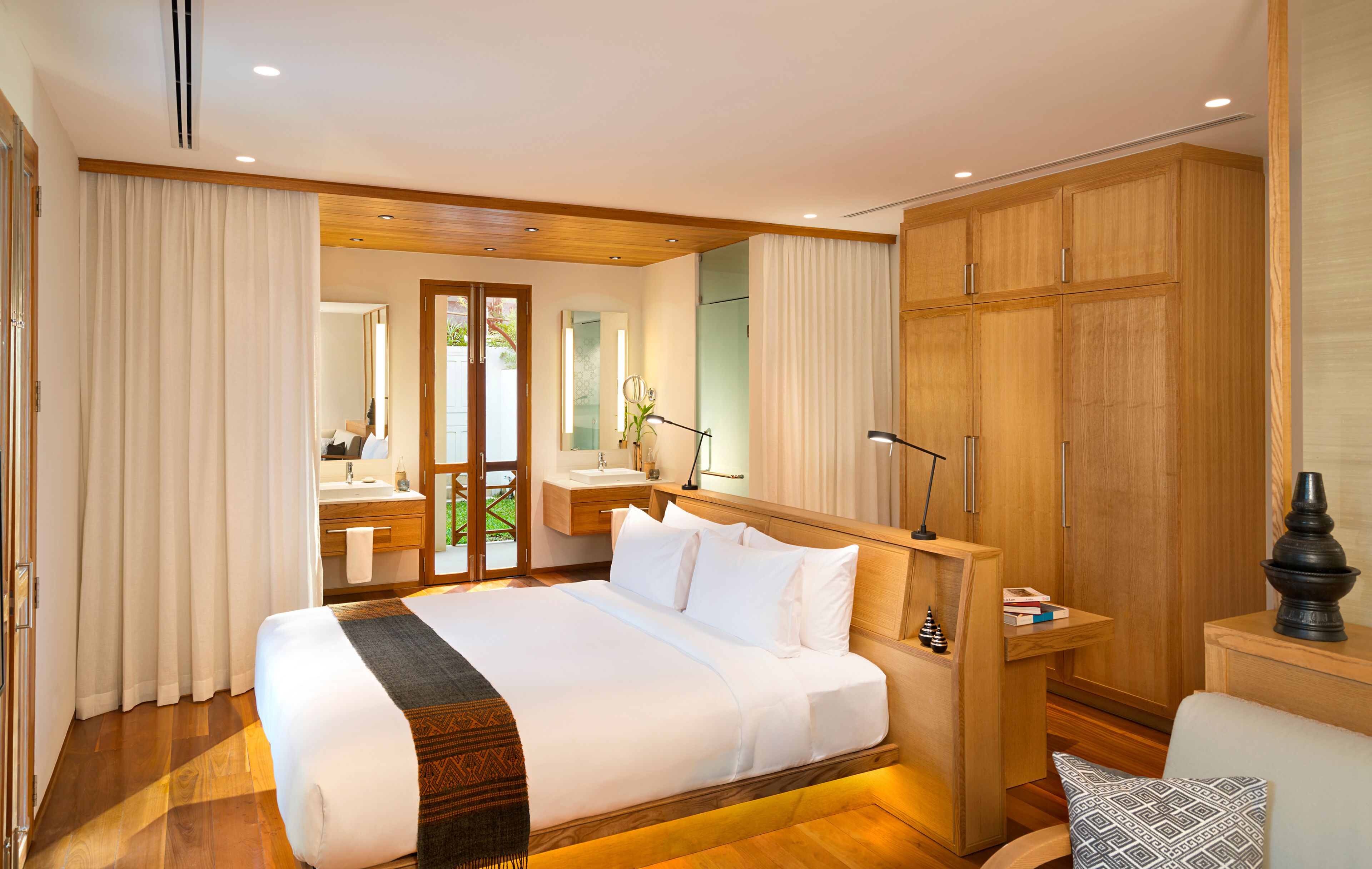 Quarto de hotel com cama grande, guarda-roupa de madeira, casa de banho com lavatório e espelho, e vista através de uma porta de vidro.