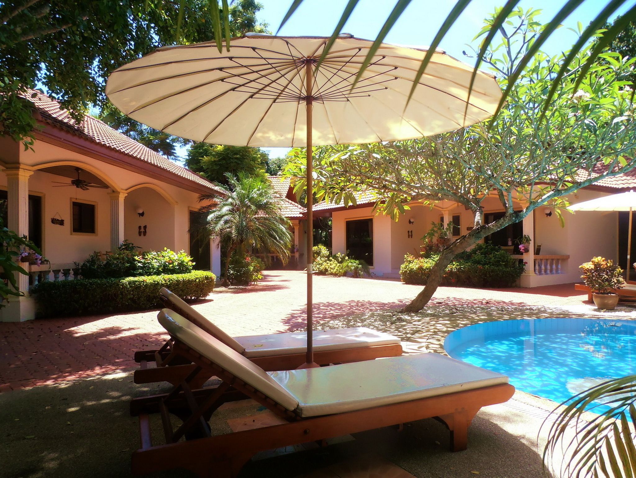 A poolside area with lounge chairs and an umbrella, surrounded by a garden and a building with a tiled roof.