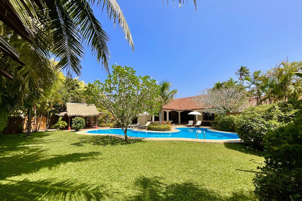 A swimming pool surrounded by a well-maintained lawn, with a thatched-roof structure and a covered patio area.