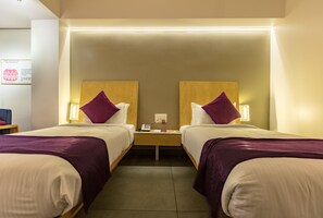 A hotel room with two beds, each with a wooden headboard, white bedding, and purple throw pillows. There is a nightstand between the beds with a phone and a lamp.