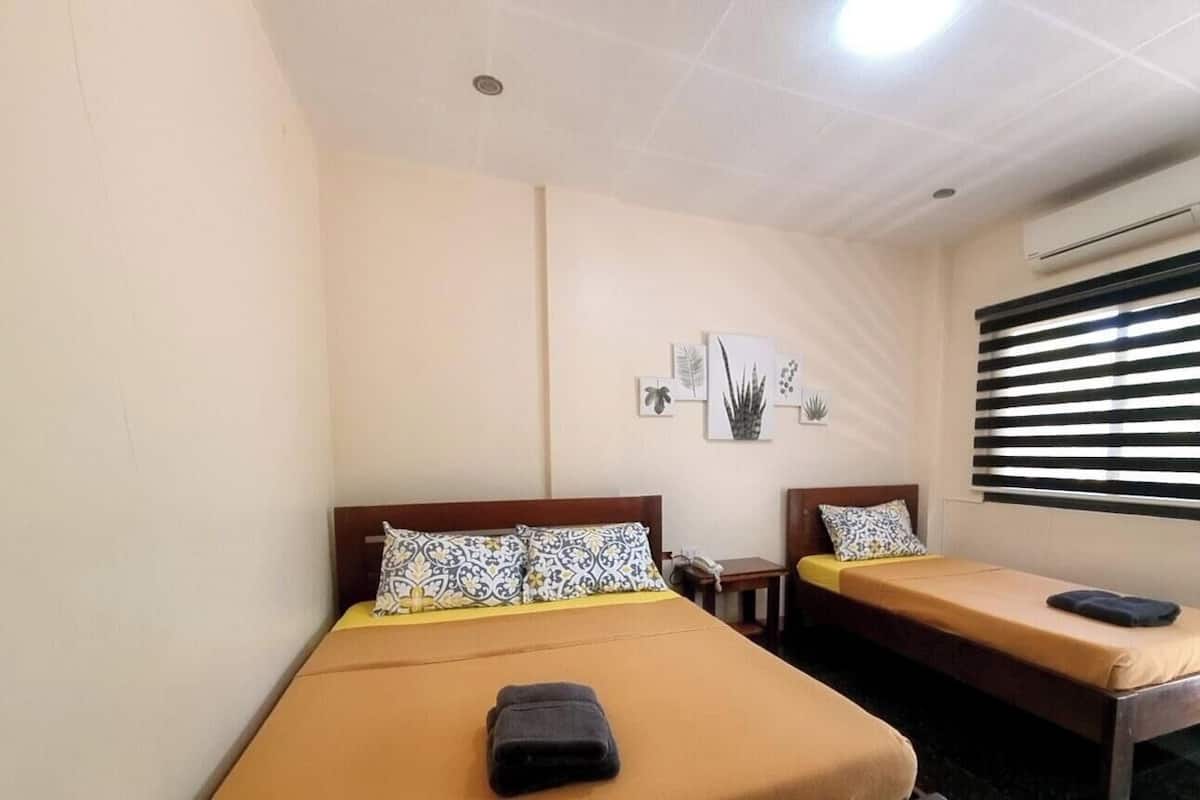 A room with two beds, a small table, a wall-mounted air conditioner, and a window with blinds.