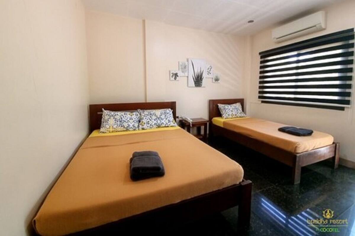 A hotel room with two single beds, a small bedside table with a lamp, a wall-mounted air conditioner, and a window with blinds.