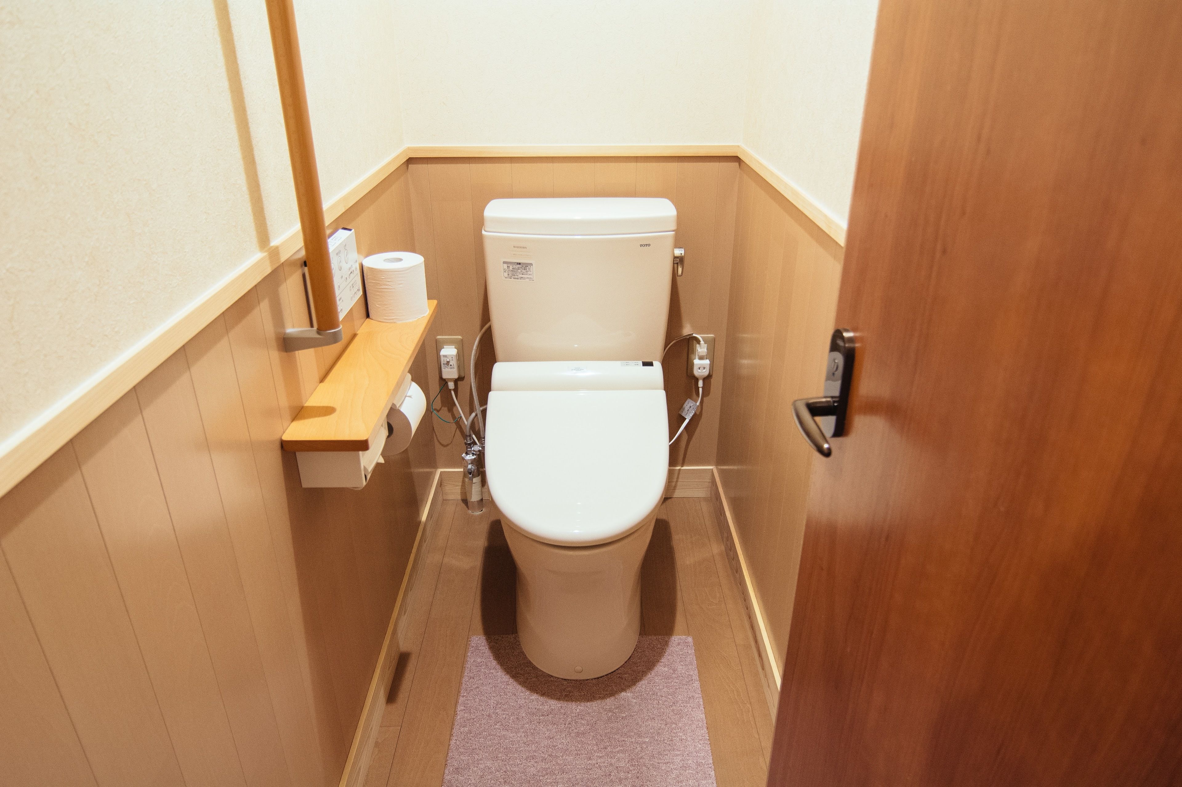 Un baño pequeño y funcional con inodoro, un estante de madera con papel higiénico y una puerta de madera.
