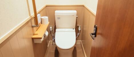 Un baño pequeño y funcional con inodoro, un estante de madera con papel higiénico y una puerta de madera.