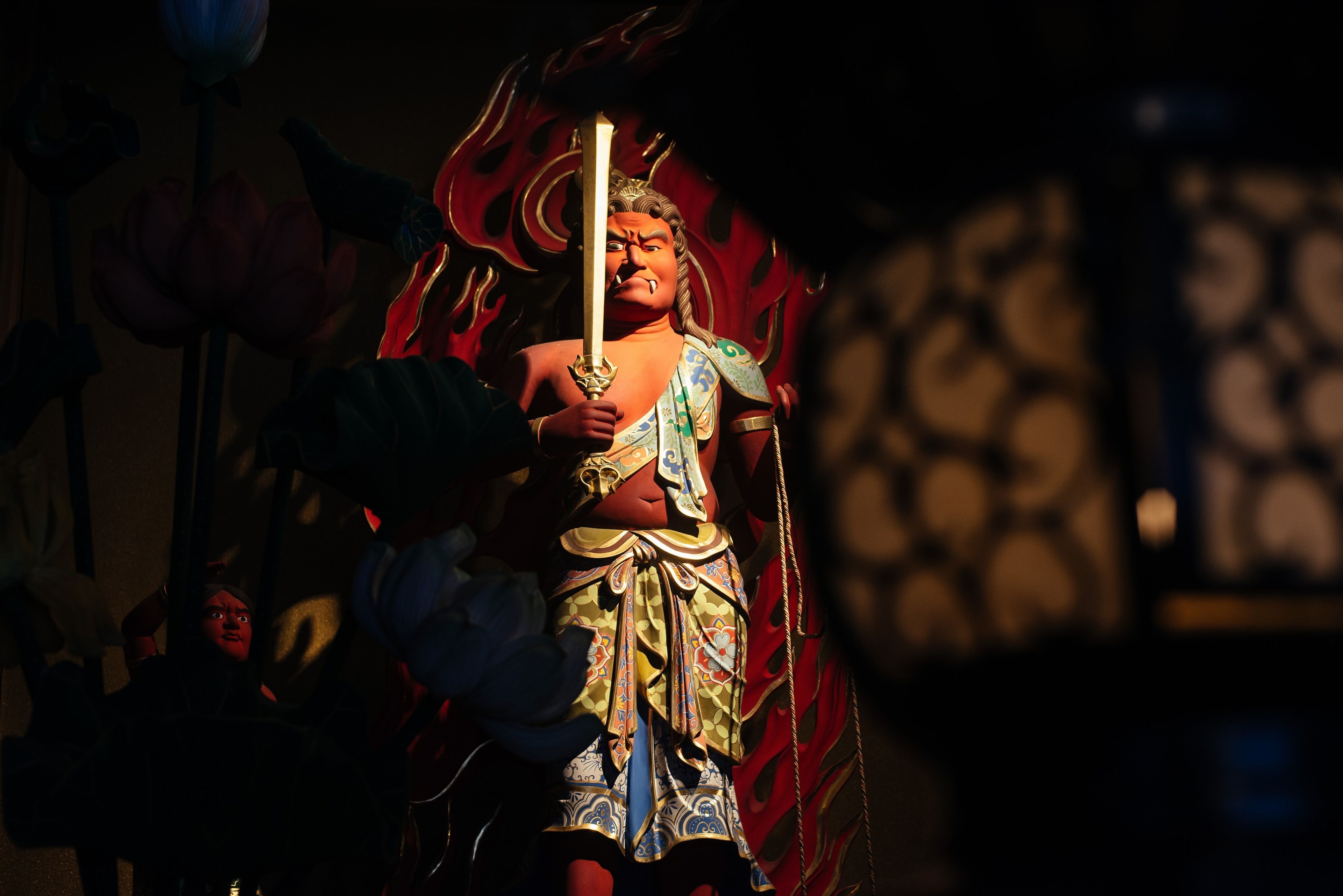 A person in traditional costume with a sword, standing in a dimly lit room with patterned walls.