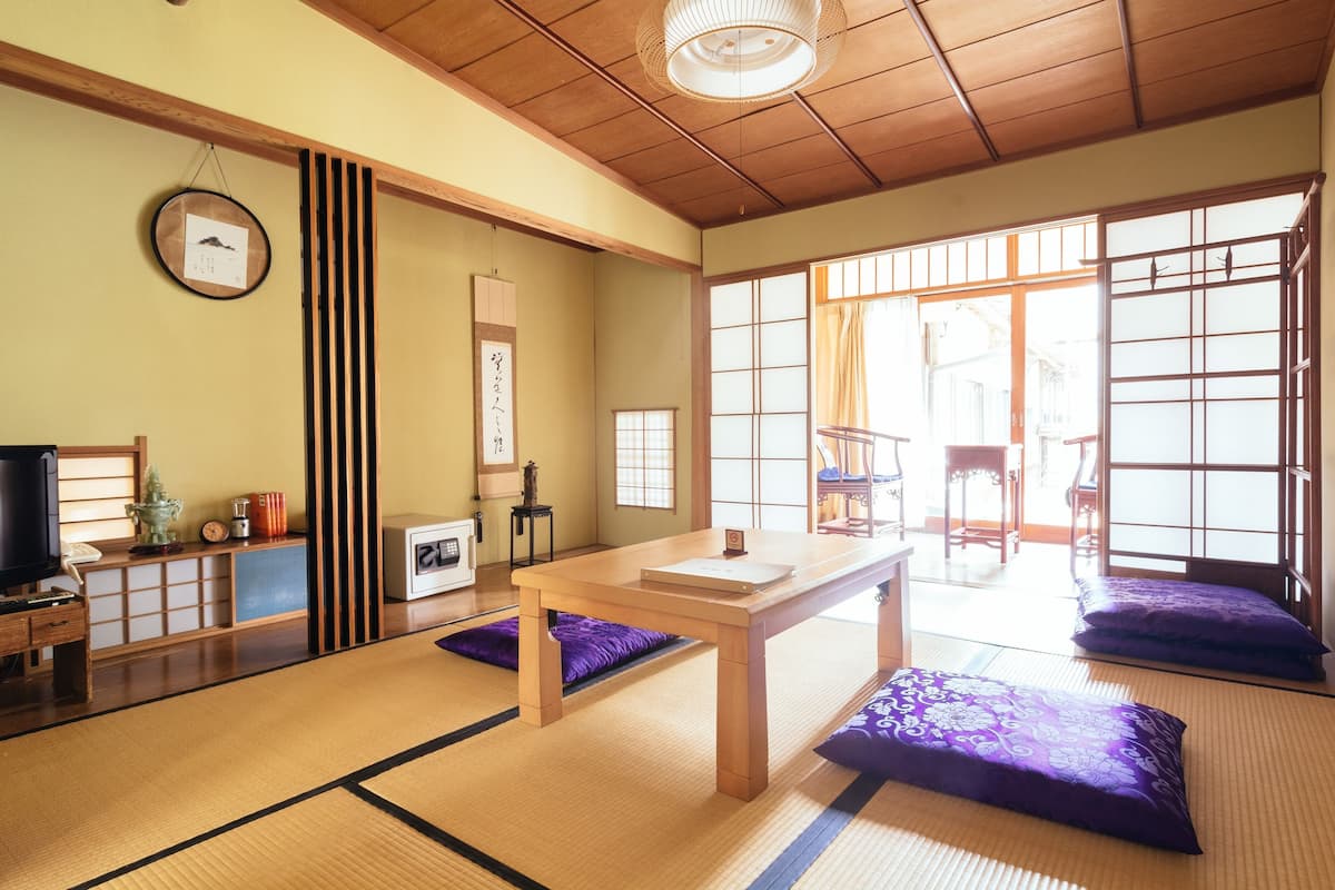 Una habitación japonesa tradicional con suelo de tatami, una mesa de madera, cojines morados, puertas correderas y un televisor.