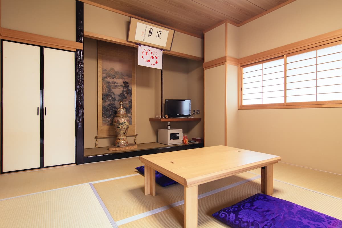 Una habitación tradicional japonesa con suelo de tatami, una mesa de madera baja, puertas correderas, un estante con televisor y una ventana con celosías shoji.