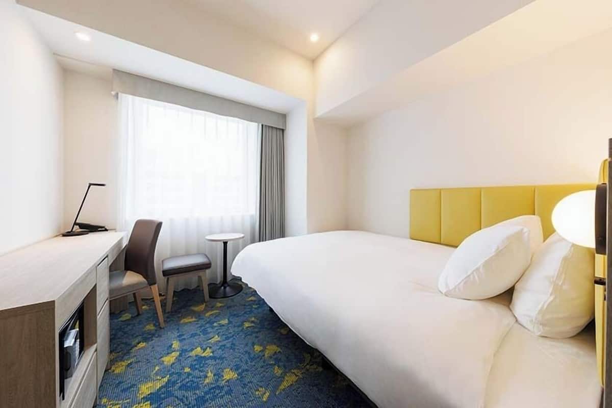 Ein Hotelzimmer mit Bett, Schreibtisch, Stuhl, einem kleinen Tisch und einem Fenster mit Vorhängen.