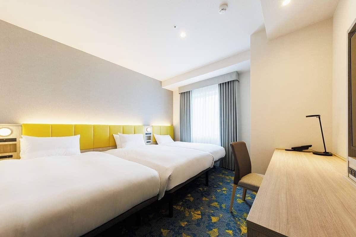 Ein Hotelzimmer mit zwei Betten, einem Schreibtisch, einem Stuhl und einem großen Fenster mit Vorhängen.
