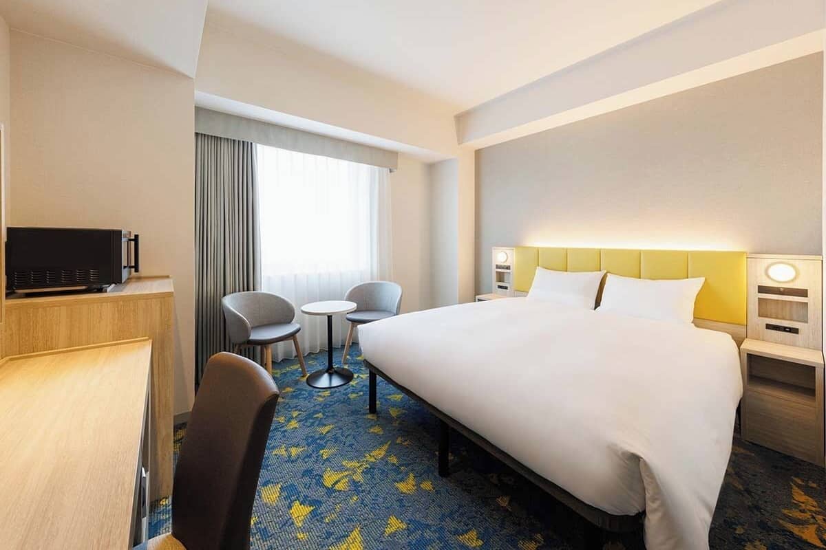 Ein Hotelzimmer mit einem großen Bett, einem Schreibtisch mit Fernseher, zwei Stühlen, einem kleinen Tisch und einem Fenster mit Vorhängen.