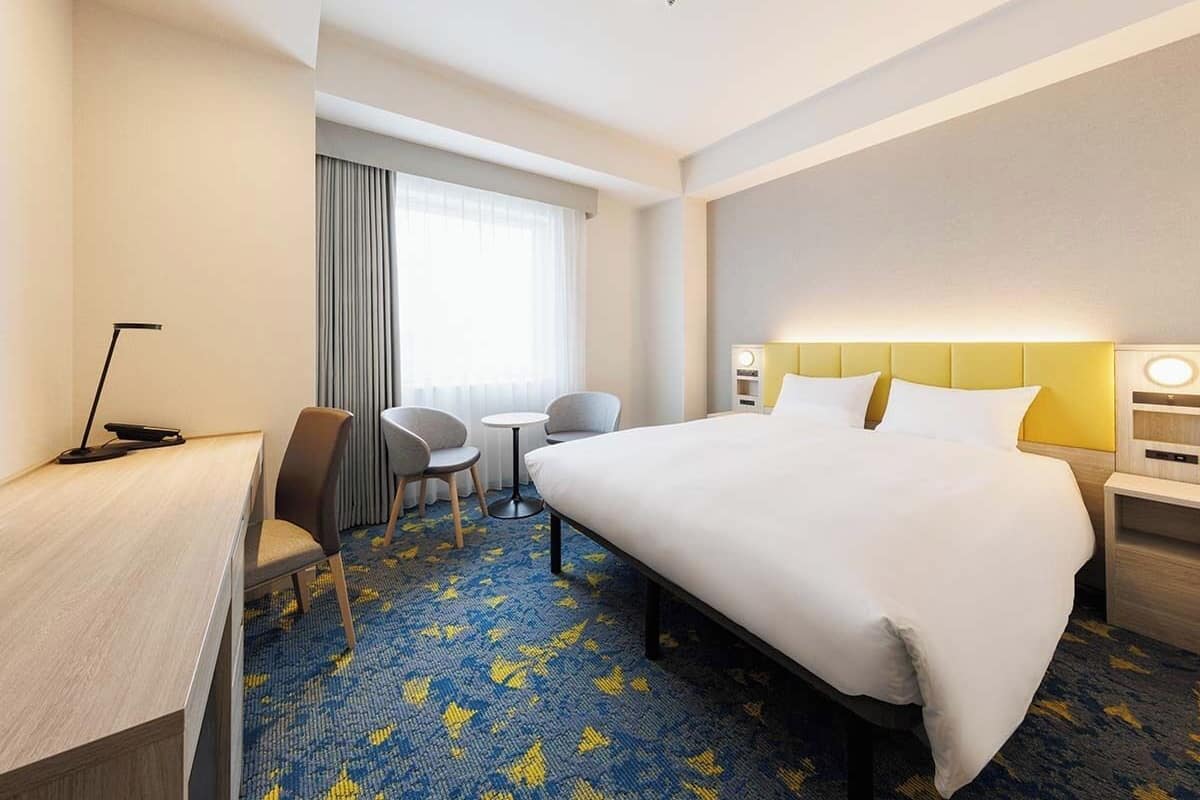 Ein Hotelzimmer mit einem großen Bett, einem Schreibtisch mit Stuhl, einem kleinen Tisch für zwei Personen, einem Fenster mit Vorhängen und einem Kopfteil mit Lampe.