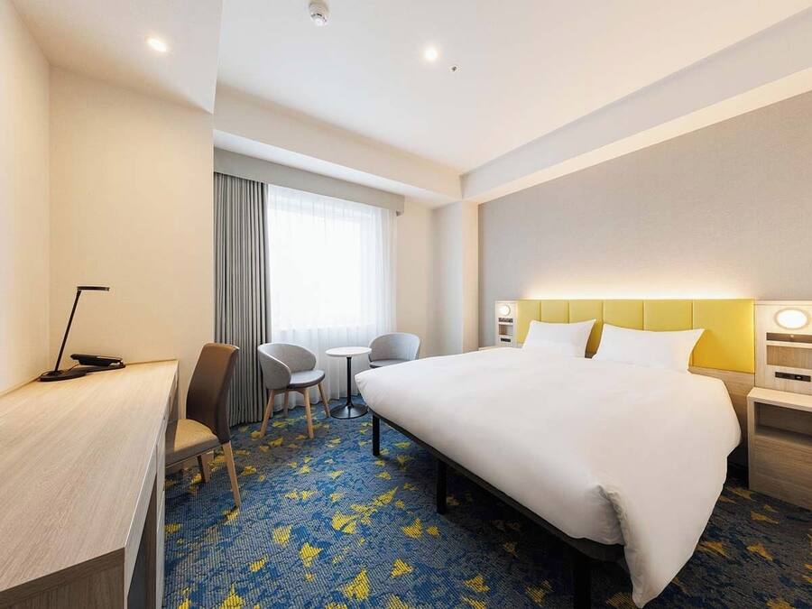 A hotel room with a large bed, a desk with a chair, a small table with two chairs, a window with curtains, and a headboard with a light.