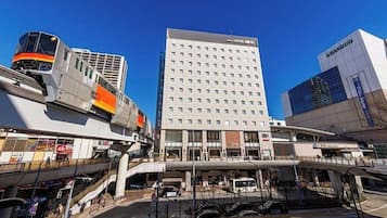 Un hotel moderno con un llamativo letrero Grandduo, un edificio de varios pisos con fachada plana, y un andén de estación de tren con un tren y un pasillo cubierto.