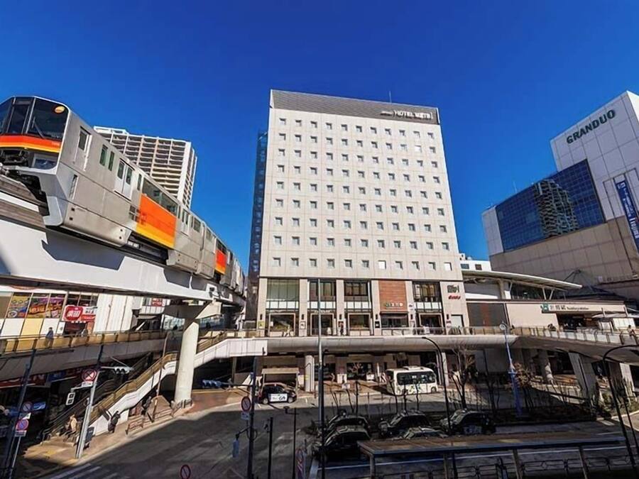 Un hotel moderno con un llamativo letrero Grandduo, un edificio de varios pisos con fachada plana, y un andén de estación de tren con un tren y un pasillo cubierto.