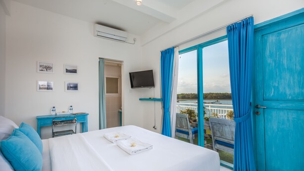 A hotel room with a bed, a desk, a TV, and a balcony with blue curtains. - Paras Paros Marina Lodge (Serangan)