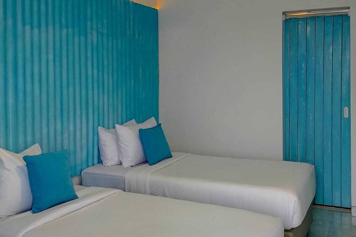 A hotel room with two beds, a blue accent wall, a ceiling light, and a door with a blue panel.
