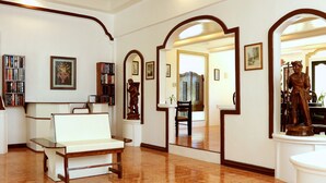 A spacious living room with a white sofa, a wooden bench, and a bookshelf filled with books. There are multiple framed pictures on the walls and a statue on a pedestal.