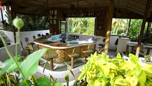 A tropical-themed bar area with a curved wooden counter, multiple seating options, and a view of lush greenery outside.