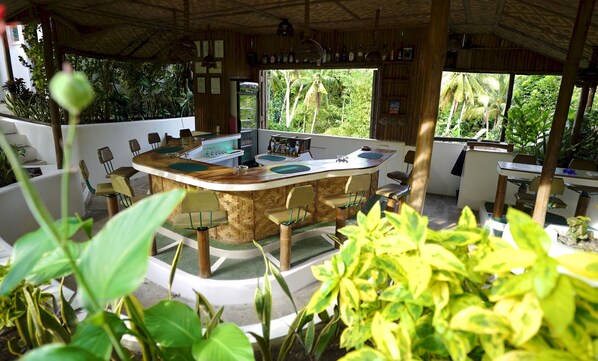 A tropical-themed bar area with a curved wooden counter, multiple seating options, and a view of lush greenery outside.