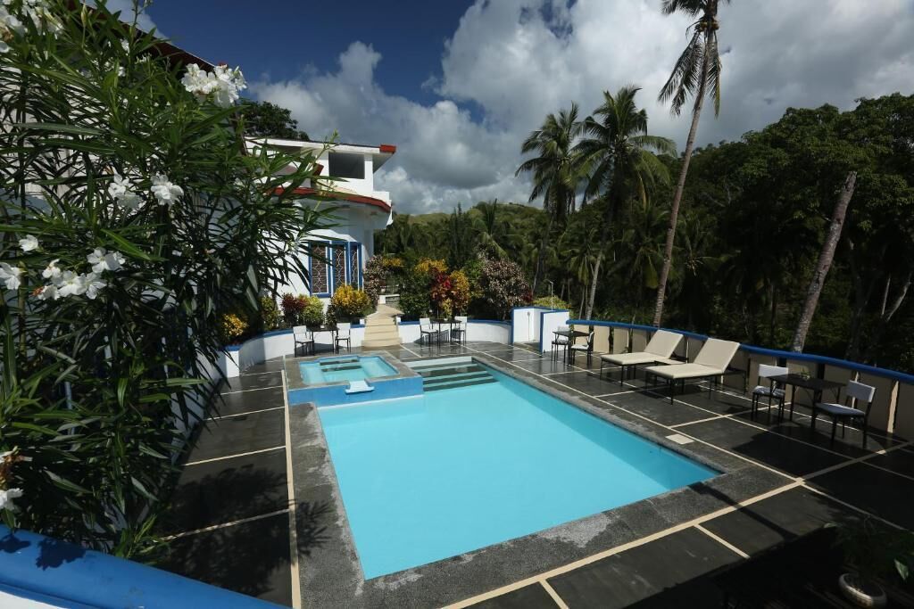 A swimming pool with lounge chairs and a small waterfall feature, surrounded by lush greenery and a clear sky.