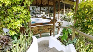 A covered outdoor seating area with a wooden table and chairs, surrounded by lush greenery.