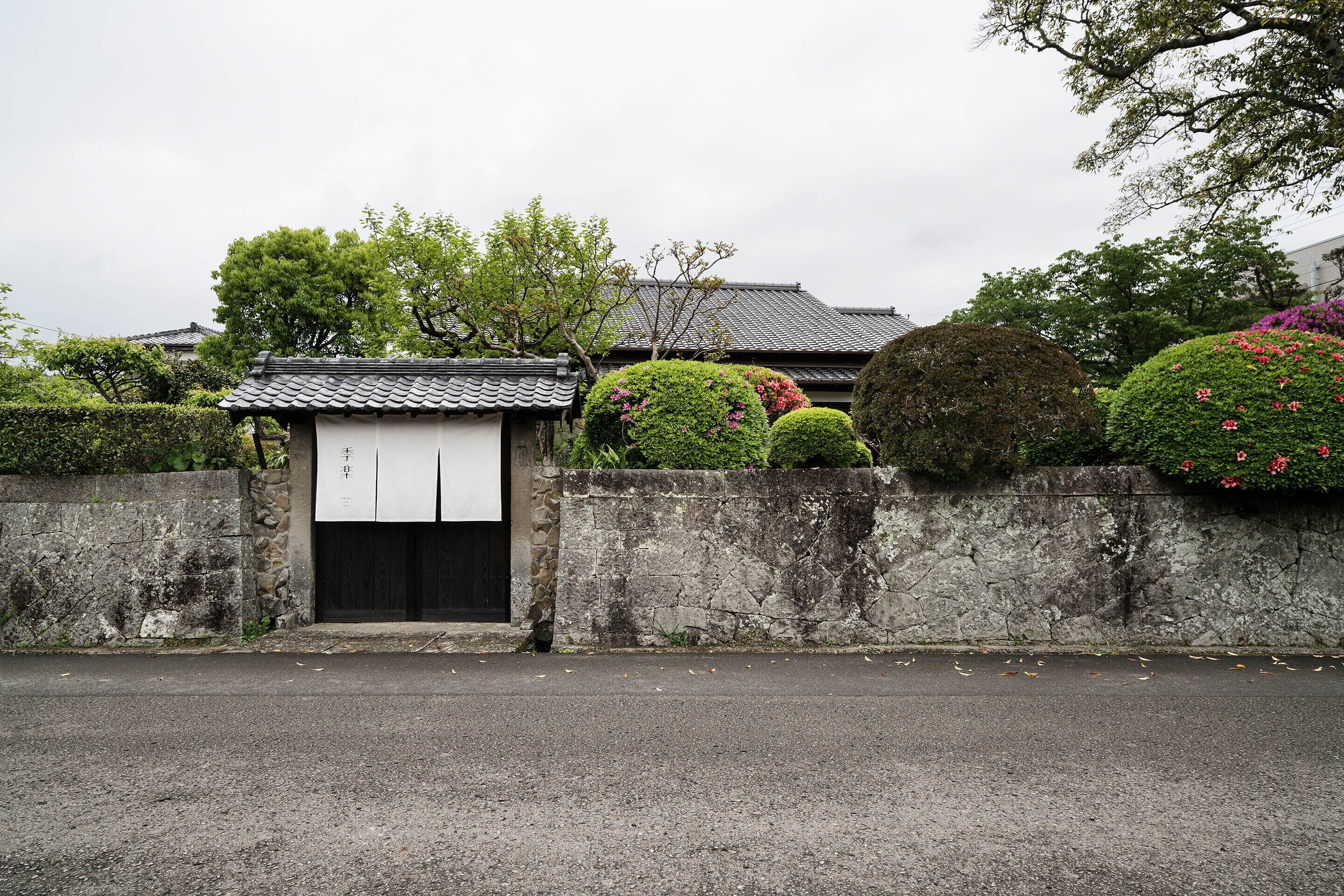 A traditional Japanese gate with a stone wall, a tiled roof, and well-maintained bushes and flowers.