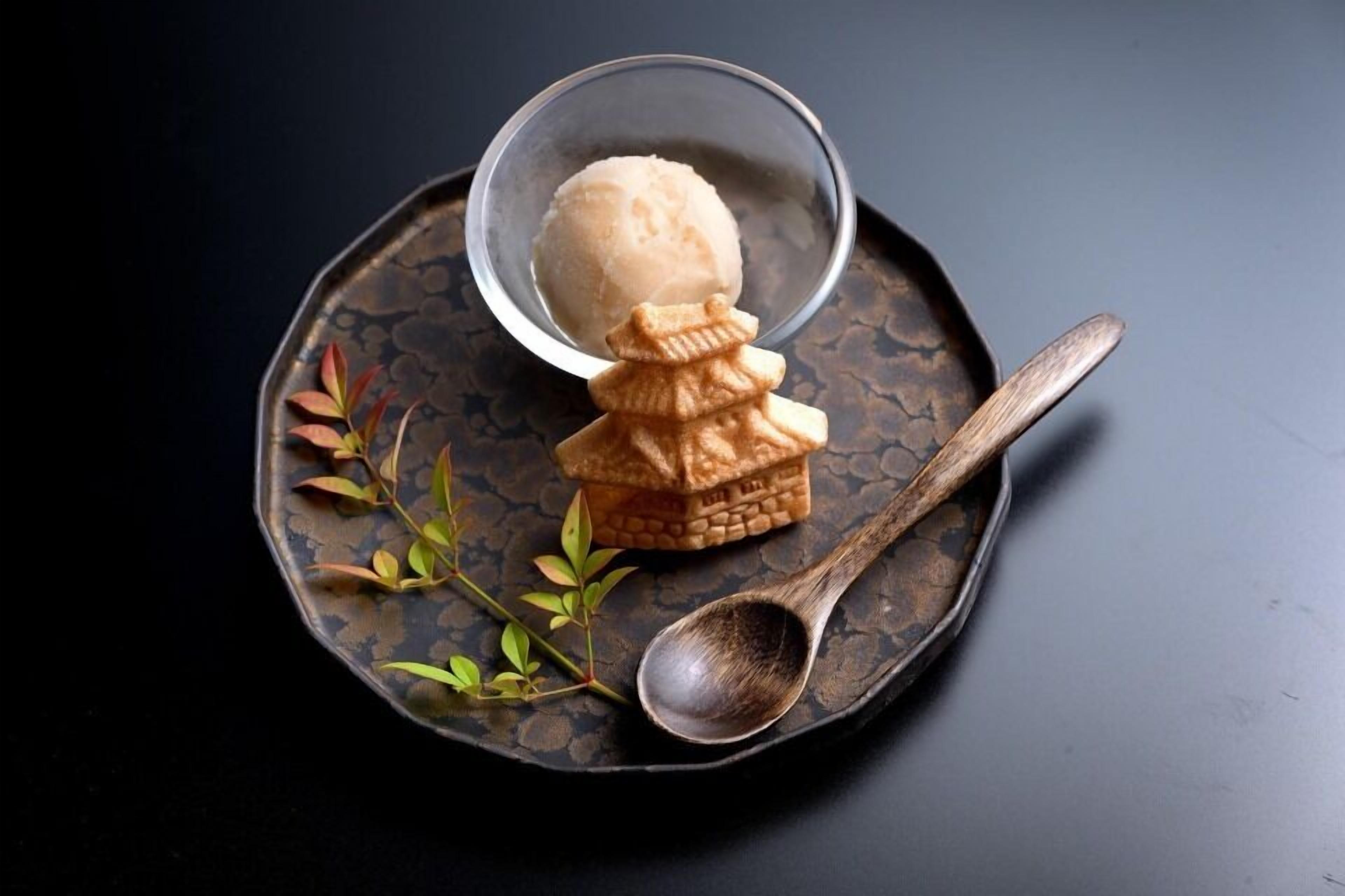 A dessert with a small house-shaped pastry and a scoop of ice cream on a dark plate with a spoon.