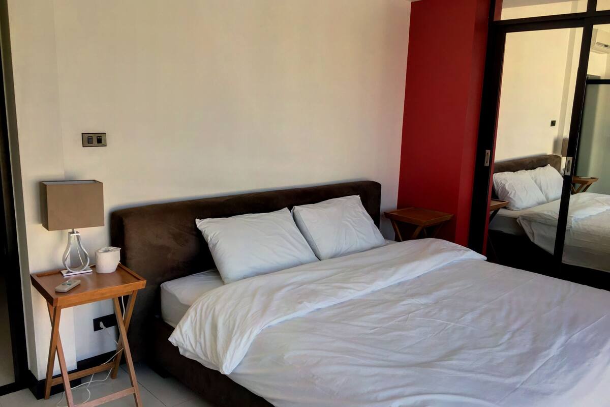 A neatly made bed with white linens, a wooden bedside table with a lamp, and a red accent wall.