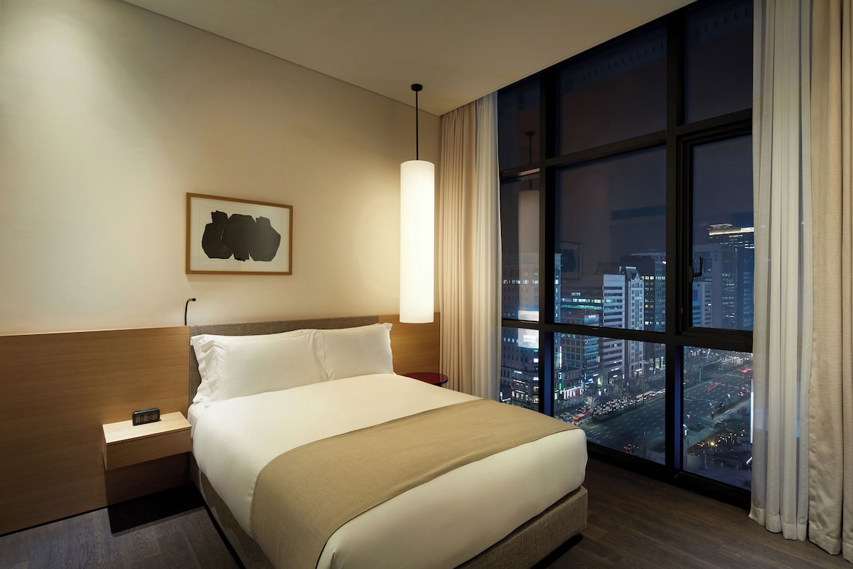 A hotel room with a large window offering a city view, a bed with white linens, a bedside table with a lamp, and a framed artwork on the wall.