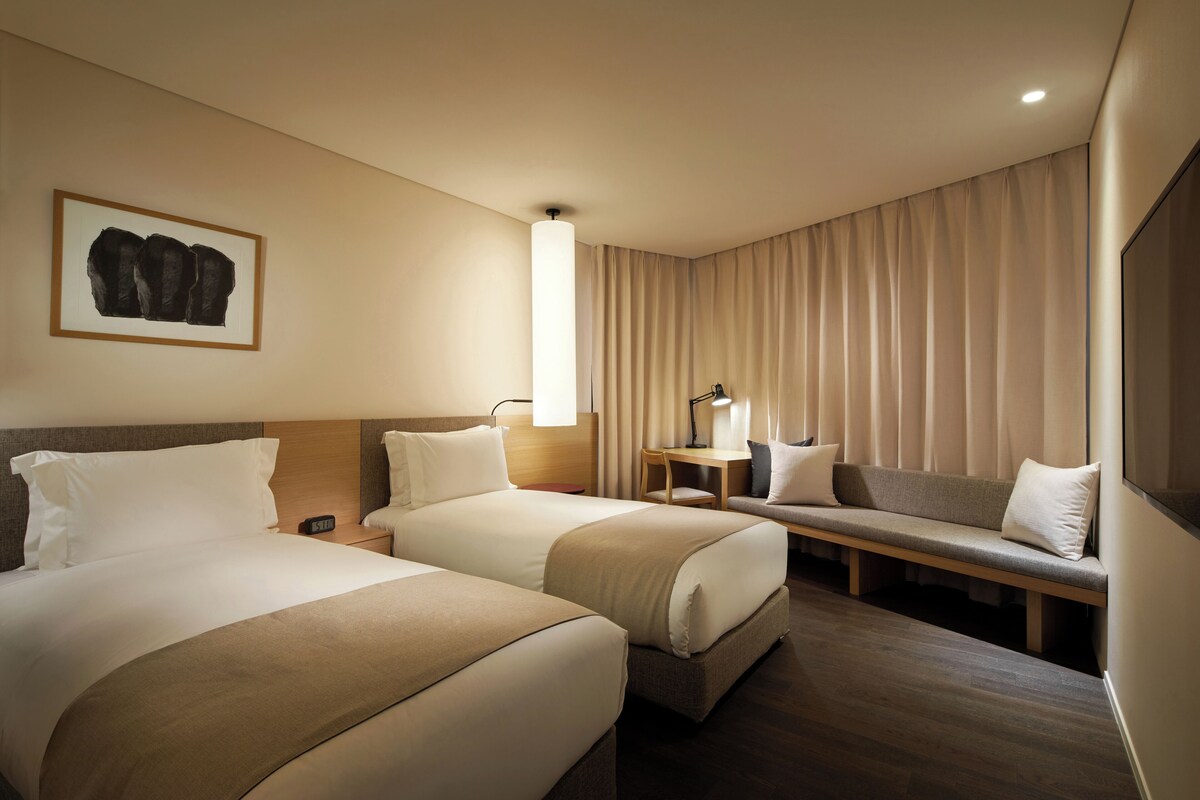 A hotel room with two beds, a desk, a bench, and a large window with curtains.