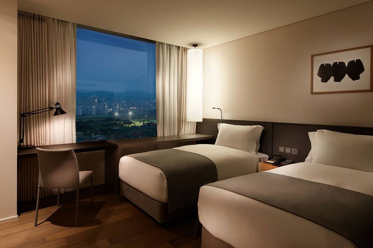 Habitación de hotel con dos camas, un escritorio, una silla y un amplio ventanal con vistas a la ciudad.
