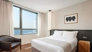 A hotel room with a large window, a bed with white bedding, a wooden nightstand, a chair, and a framed artwork on the wall.