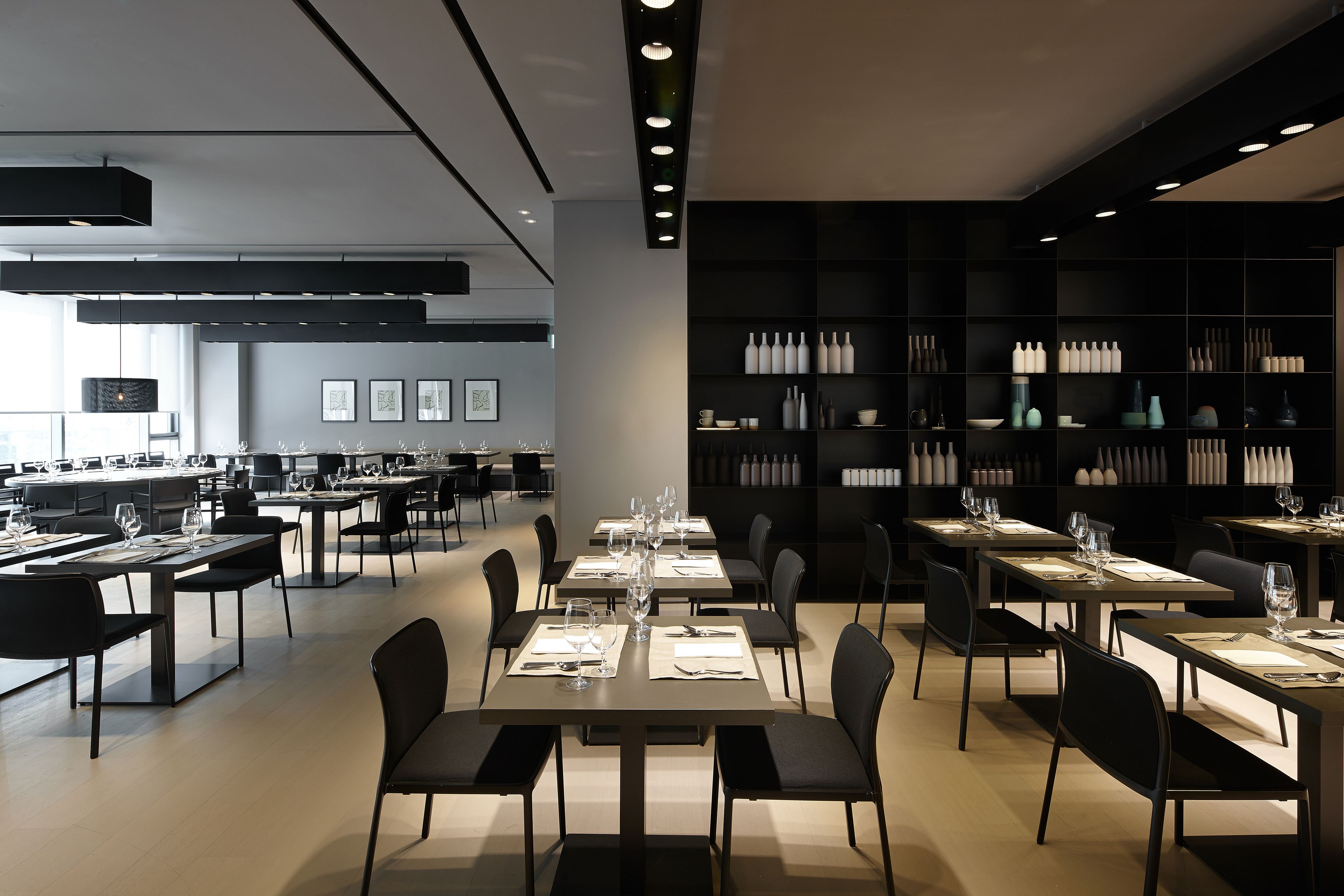 A modern dining area with multiple tables and chairs, a large black shelving unit with bottles, and framed pictures on the wall.