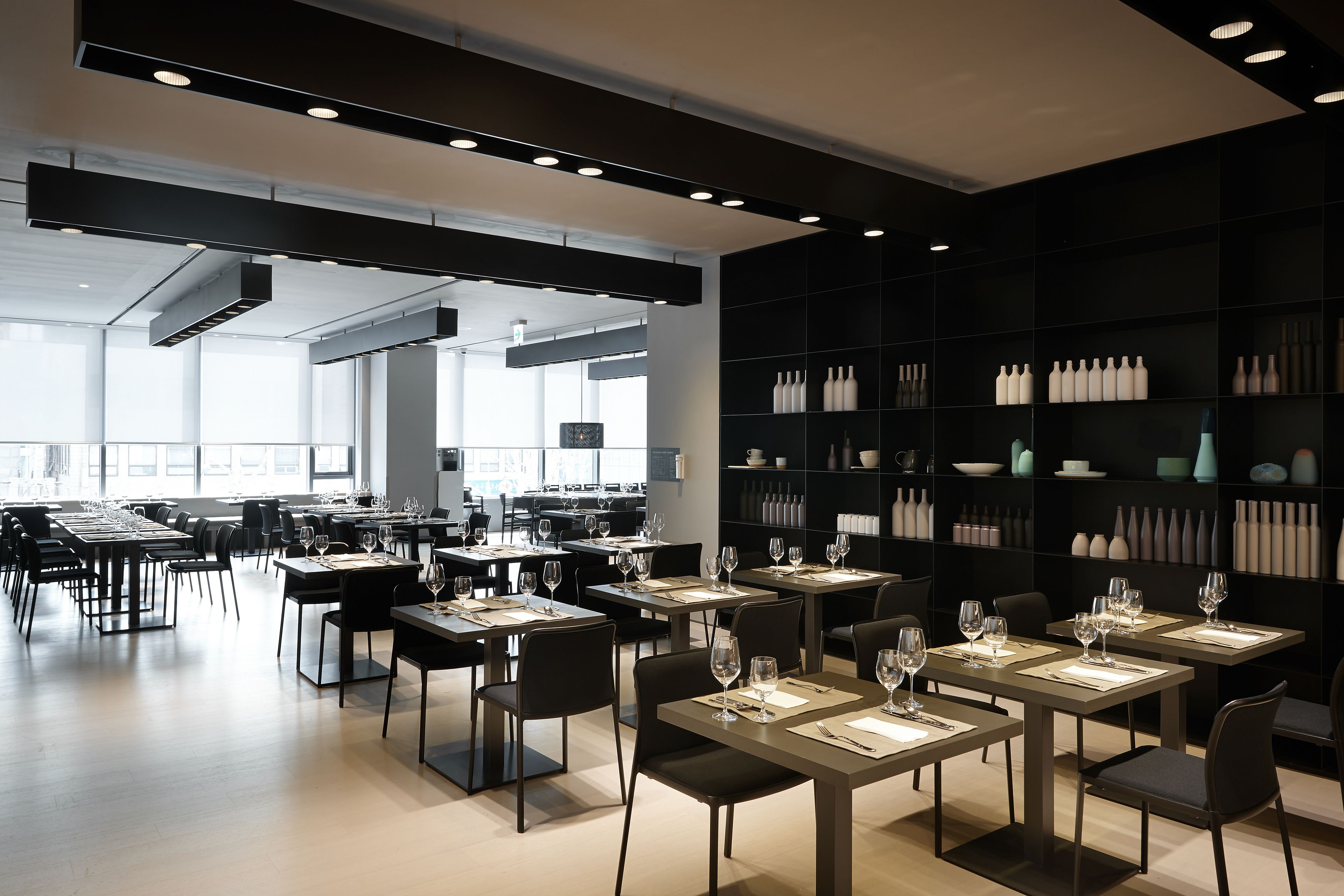 A modern dining area with tables set for service, a large black shelving unit with decorative items, and a bright, airy space with natural light.