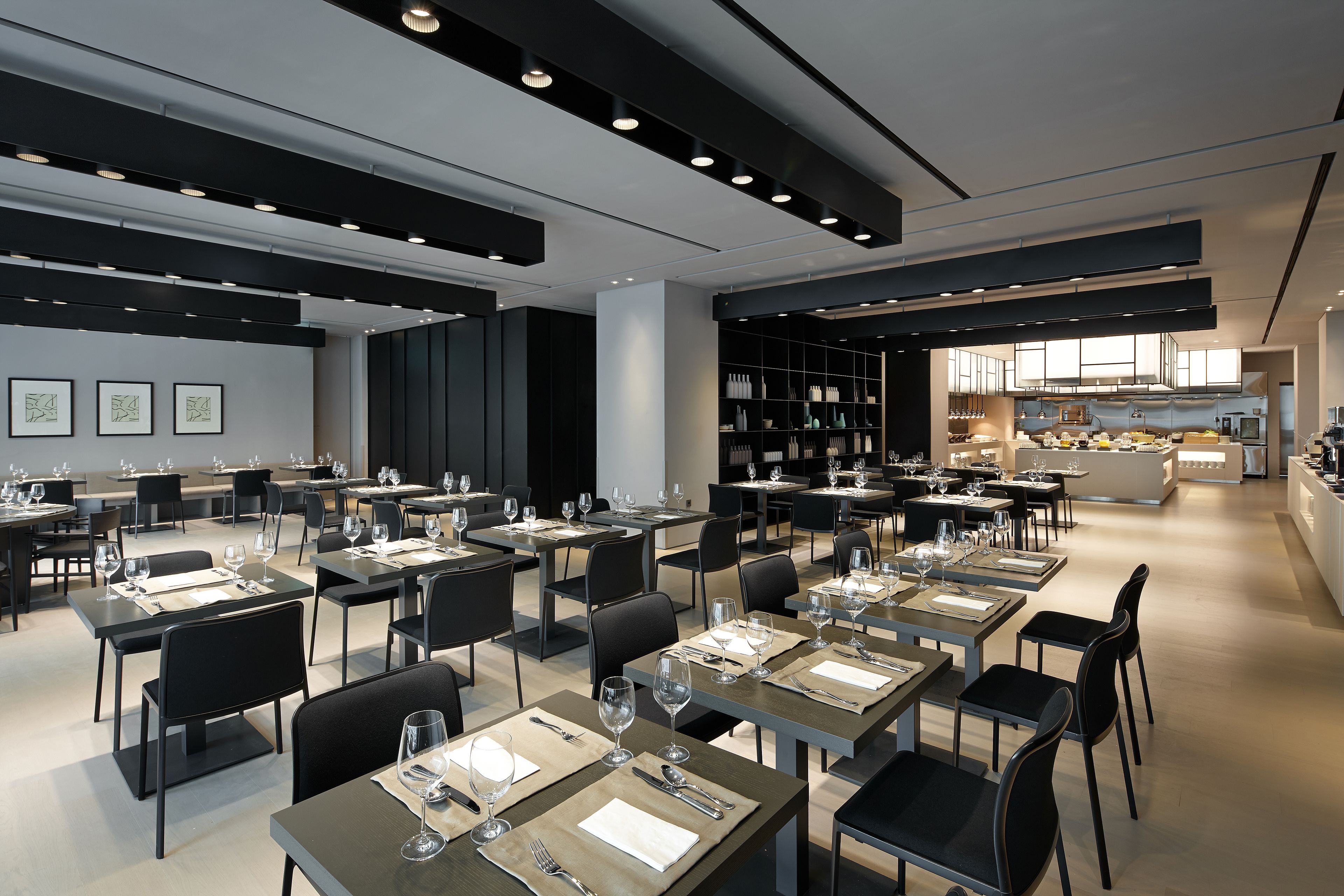 Un restaurante moderno con varias mesas y sillas, una zona de cocina al fondo y un diseño interior limpio y minimalista.