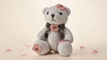 A plush teddy bear with a flower on its head and a flower on its chest, sitting on a plain surface with scattered petals.