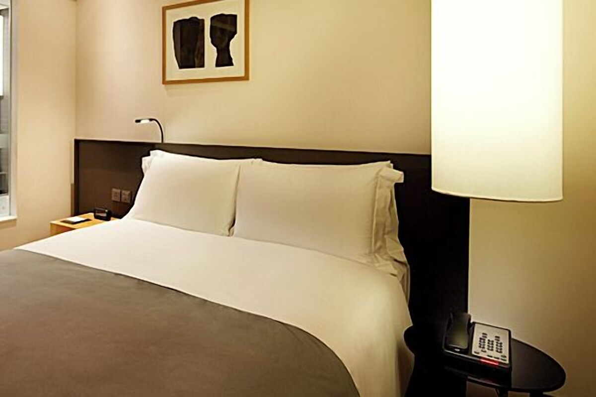 A hotel room with a large bed, a bedside table with a lamp, a nightstand, a mirror, and a framed artwork on the wall.